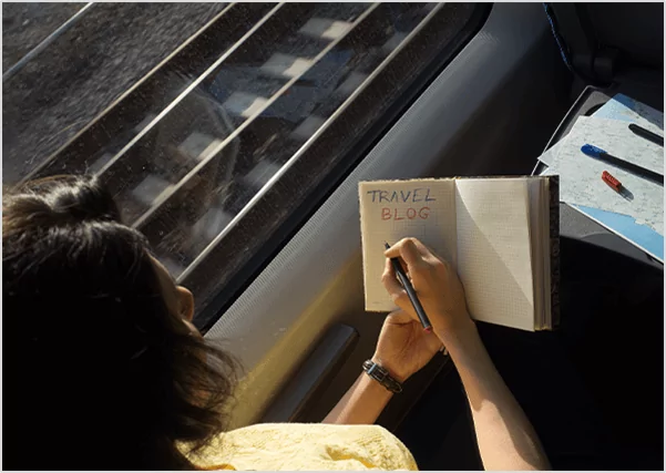 A woman writing "TRAVEL BLOG" in a notebook while looking out a train window, illustrating professional travel writing and blog publishing services.