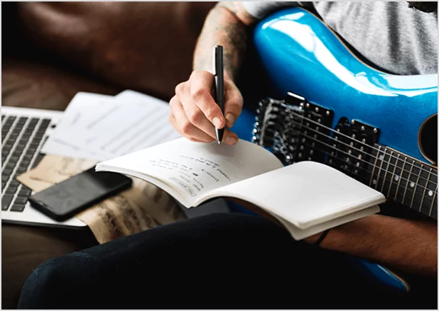 Close-up of a person’s hands writing lyrics in a notebook while holding guitar, representing creative songwriting workshops