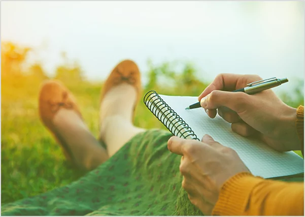 Close-up of a person writing in a spiral notebook while relaxing in a grassy field, illustrating creative poetry and prose writing services.