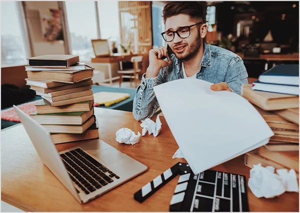 A young man with glasses sitting at a cluttered desk, talking on a smartphone while holding out a white manuscript page, surrounded by stacks of books and a film clapperboard.