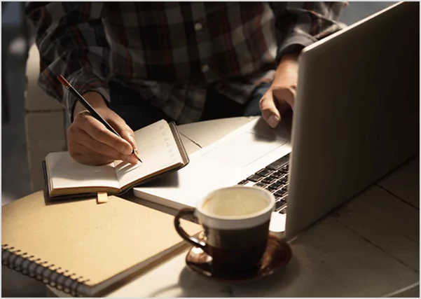 Alt Text: A person wearing a plaid shirt sitting at a desk, multitasking by writing in a small notebook with a pencil while simultaneously using a laptop.