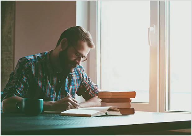 A bearded man with glasses sitting at a table by a window, focused on writing in a notebook next to a stack of books and a mug.
